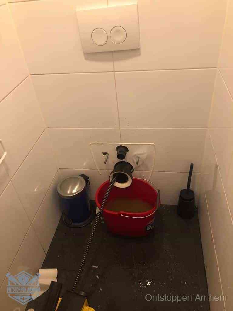 Een ontstopte wand-wc met verwijderde toiletpot waar een ontstoppingsveer in de wanduitgang verdwijnt, het achterhalen waar de verstopping zit in Arnhem