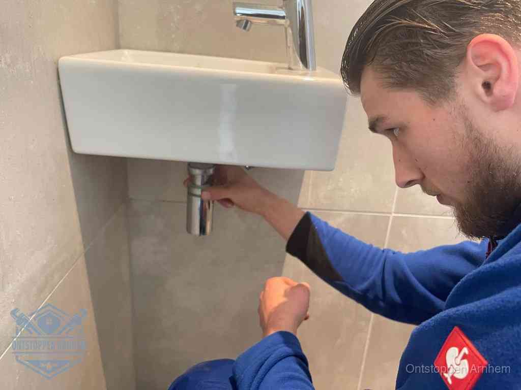 Een monteur draait met de hand de zwanenhalssifon onder een fonteinwasbak in de toiletruimte los om de bron van rioollucht in Arnhem te vinden