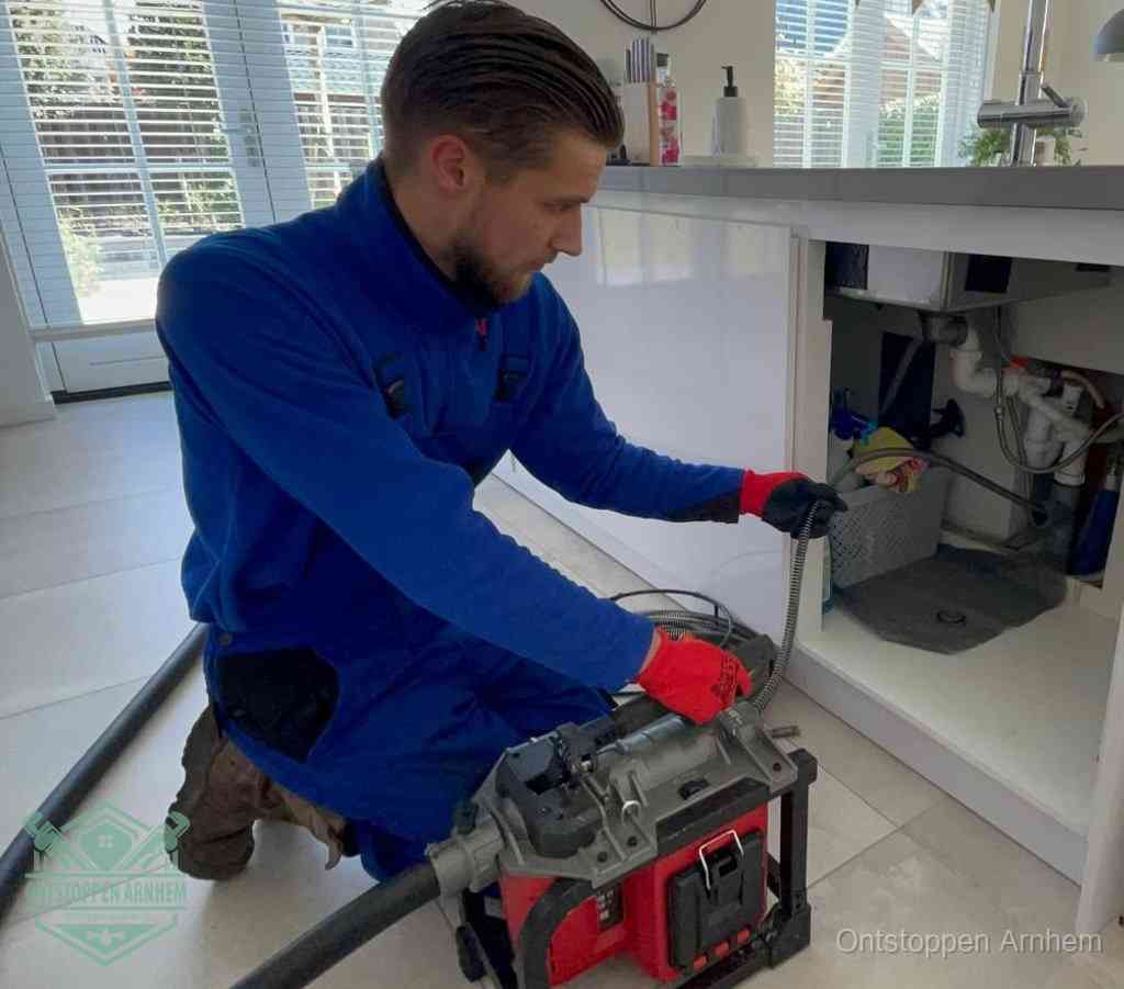 Een monteur met rode handschoenen knielt in een keuken en voert een elektrische ontstoppingsveer in tijdens het professioneel afvoer ontstoppen in Arnhem